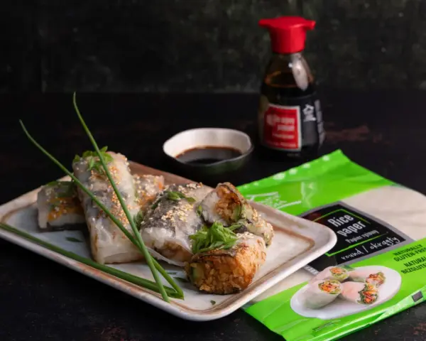 Pyszne sushi w papierze ryżowym, posypane sezamem, podane z sosem sojowym. Obok opakowanie papieru ryżowego.