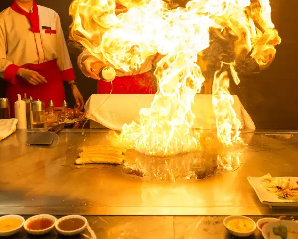 Pokaz teppanyaki z płomieniami buchającymi z płyty grillowej. Kucharz w czerwonym fartuchu dodaje składniki, tworząc widowiskowy spektakl.