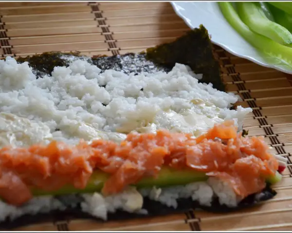 Przygotowanie hosomaki oshinko: ryż, łosoś i ogórek na nori, gotowe do zwinięcia.