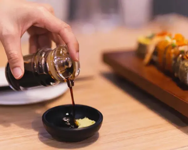 Dłoń nalewa sos sojowy bezglutenowy do miseczki z wasabi. Obok leży sushi.
