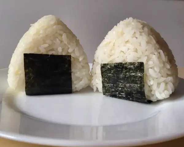 Dwa pyszne onigiri sushi, uformowane w trójkąty i ozdobione kawałkami nori, leżą na białym talerzu.