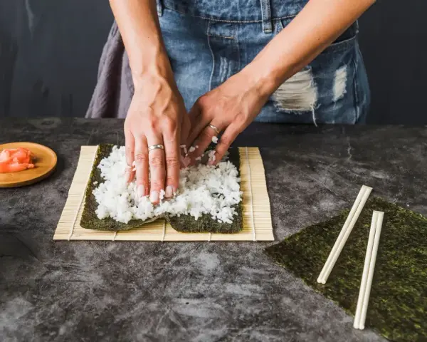 Ręce rozkładają ryż na nori, przygotowując się do zwinięcia sushi. Obok leżą pałeczki i talerzyk z imbirem, idealny dodatek do sushi.