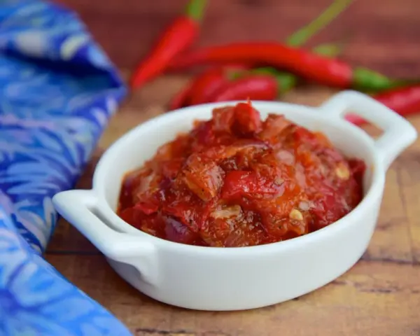 Pikantny sos sambal oelek w białej miseczce, obok czerwonych papryczek chili i niebieskiej tkaniny.