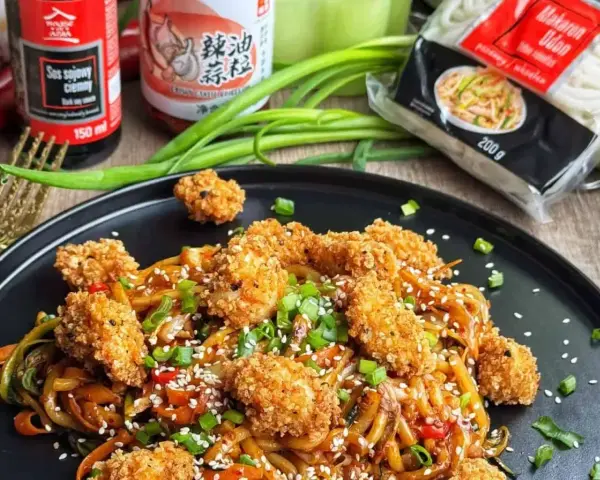 Pyszny **udon z kurczakiem i warzywami**, posypany sezamem i szczypiorkiem. Obok sosy i opakowanie makaronu.