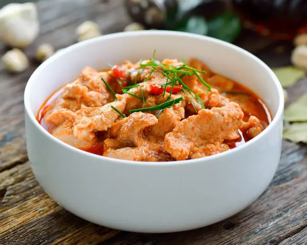 Aromaticzne curry z wieprzowiną w białej misce, ozdobione zieloną szczypiorkiem i czerwonymi papryczkami chili.