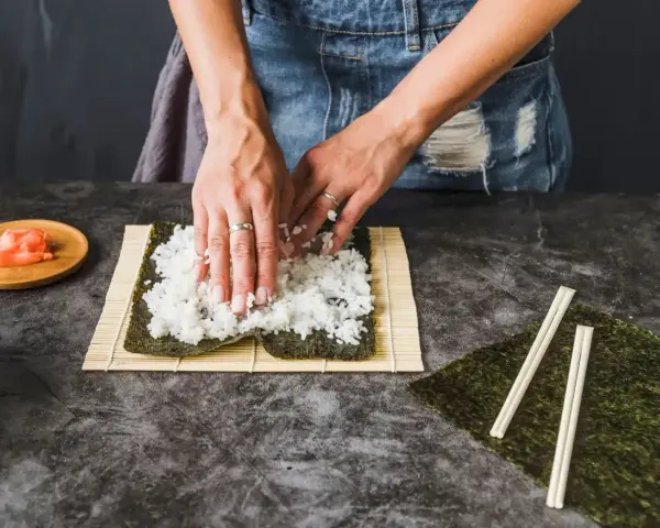 Dłonie rozprowadzają ryż na nori. Obok leżą pałeczki i marynowany imbir. To klucz do idealnego ryżu do sushi, który potrzebuje odrobiny octu.