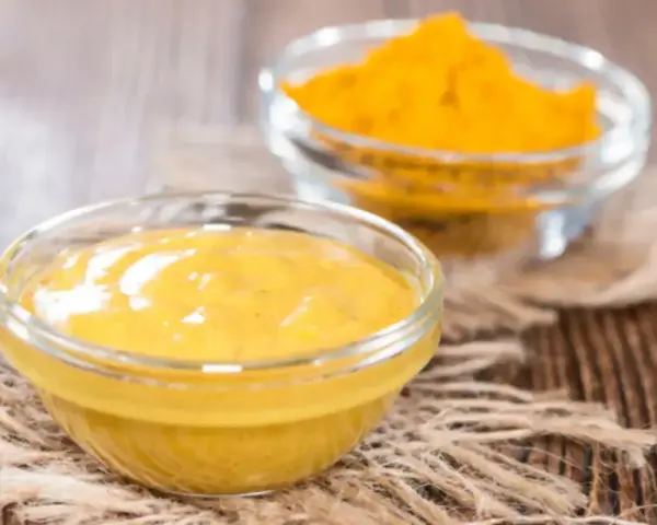 Dwie szklane miseczki na drewnianym tle. Jedna zawiera gładki sos, druga żółtą przyprawę, składniki do korma curry.