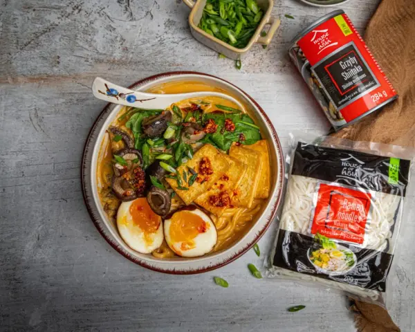 Szybki ramen przepis: miska pełna aromatycznego bulionu z grzybami shiitake, tofu, jajkiem i szczypiorkiem. Obok opakowanie makaronu i grzybów.