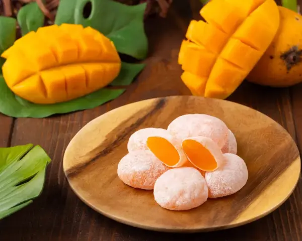 Słodkie mochi o smaku mango, ułożone na drewnianym talerzu, obok świeżych owoców i liści.