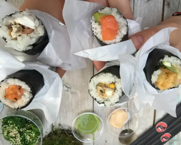 Ręce trzymają cztery pyszne temaki zawinięte w papier. Widać różne nadzienia: łososia, awokado, warzywa i rybę.
