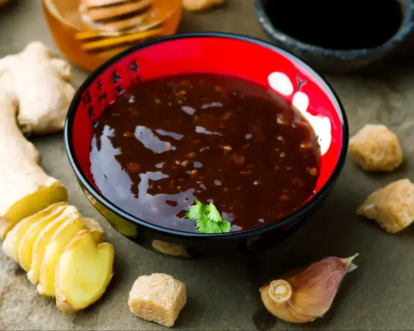Sos teriyaki przepis: miseczka z ciemnym sosem, obok imbir, czosnek, cukier trzcinowy i miód. Idealny do dań azjatyckich.
