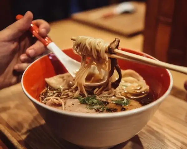 Ręka trzyma pałeczkami porcję parującego ramenu z makaronem i grzybami. Oto jak jesc ramen!