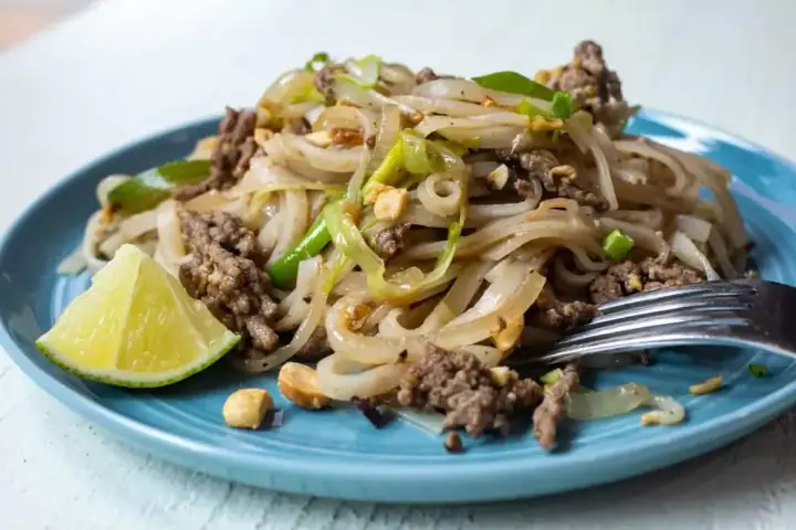 Pyszny pad thai z wołowiną, makaronem ryżowym, orzeszkami ziemnymi i ćwiartką limonki na niebieskim talerzu.