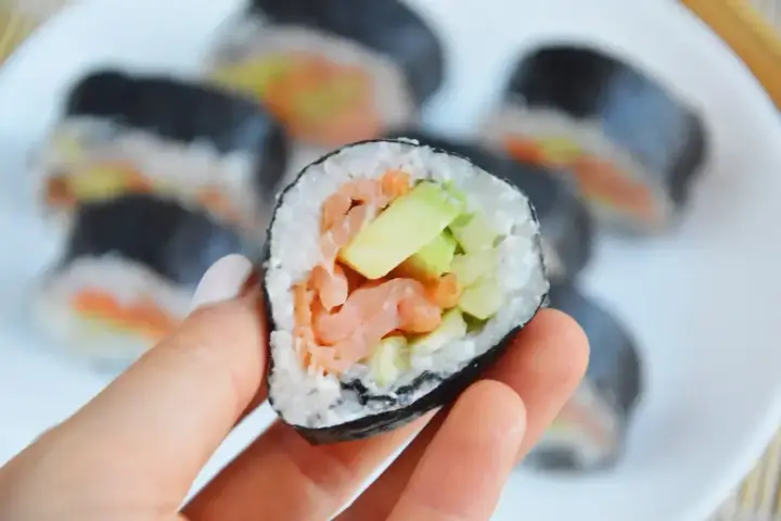 Hosomaki łosoś z awokado, trzymane w dłoni, na talerzu pełnym sushi.