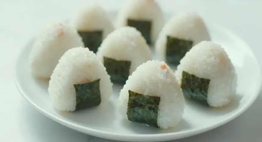 Kilka pysznych onigiri z łososiem, udekorowanych kawałkami nori, ułożonych na białym talerzu.