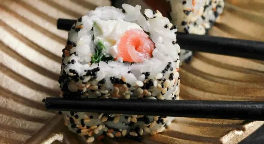 Rolka sushi z łososiem i serkiem, posypana sezamem, trzymana pałeczkami. Czy sushi można jeść na drugi dzień? Tak, jeśli było odpowiednio przechowywane.