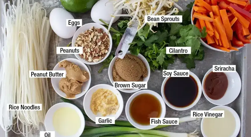 Składniki do sosu pad thai: makaron ryżowy, jajka, orzeszki ziemne, masło orzechowe, olej, czosnek, zielona cebulka, brązowy cukier, kiełki fasoli, kolendra, marchewka, papryka, sos sojowy, sos rybny, ocet ryżowy, sriracha, limonka.