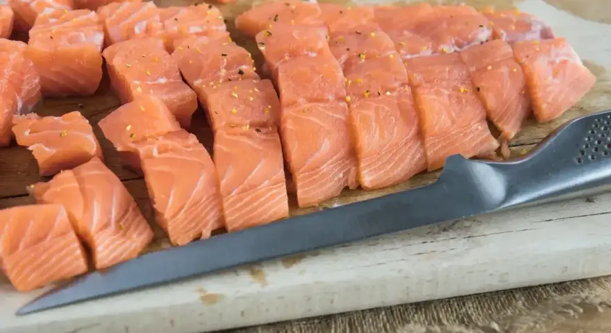 Pokrojona w kostkę i plastry świeża ryba do sushi, przyprawiona, leży na desce obok noża.