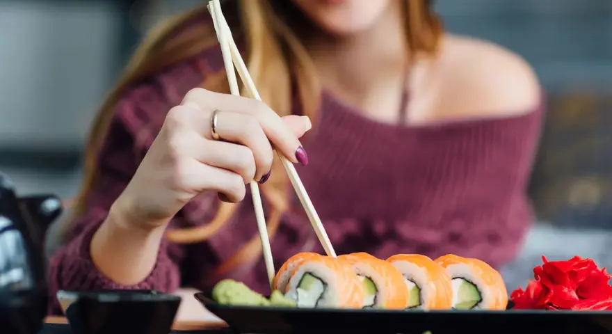Uśmiechnięta kobieta w ciąży sięga po sushi. Na talerzu rolki z łososiem i awokado, obok wasabi i marynowany imbir.