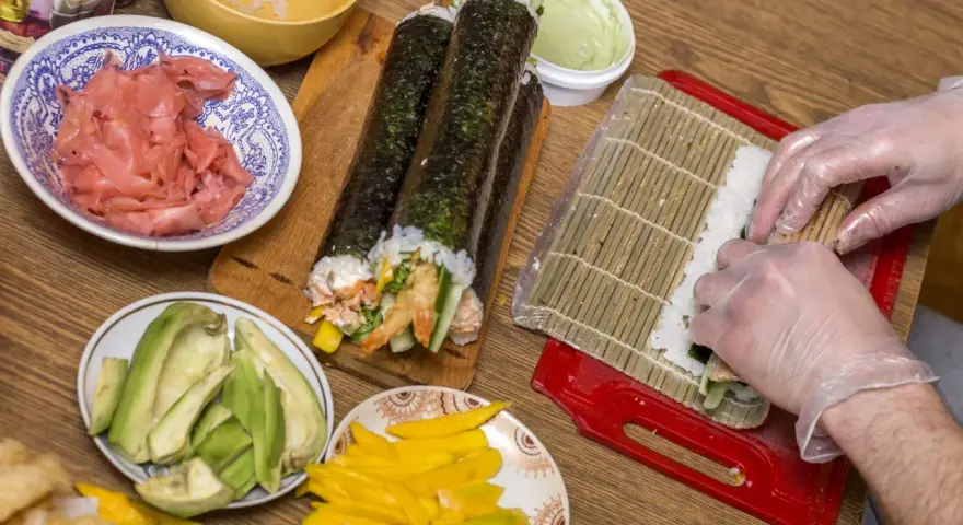 Przygotowanie sushi z warzywami: ryż, awokado, mango, krewetki i marynowany imbir. Ręce w rękawiczkach zwijają rolkę.