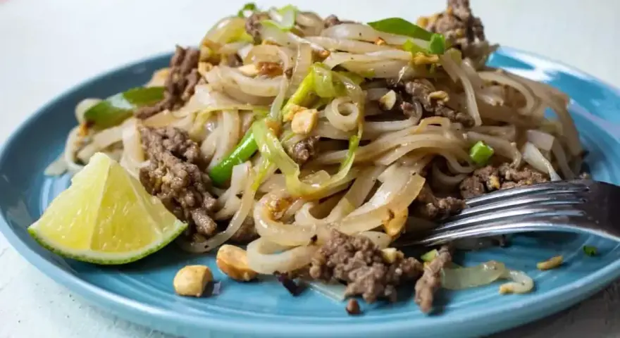 Pyszny pad thai z wołowiną, makaronem ryżowym, orzeszkami ziemnymi i ćwiartką limonki na niebieskim talerzu.
