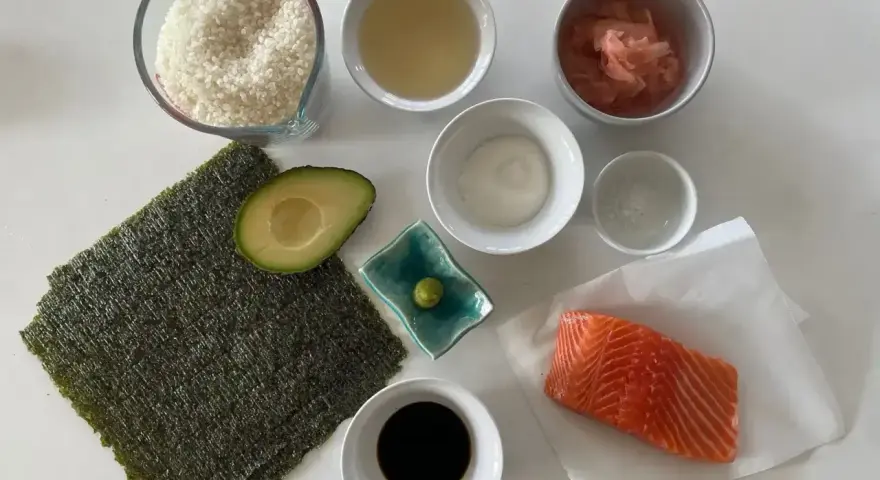 Wszystkie składniki do sushi: ryż, nori, łosoś, awokado, sos sojowy, wasabi, imbir marynowany, ocet ryżowy i sól.