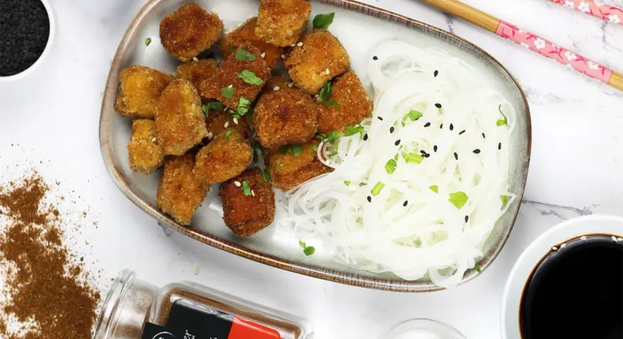 Chrupiące tofu w tempurze z makaronem ryżowym, posypane sezamem i szczypiorkiem. Obok sos sojowy i przyprawy.