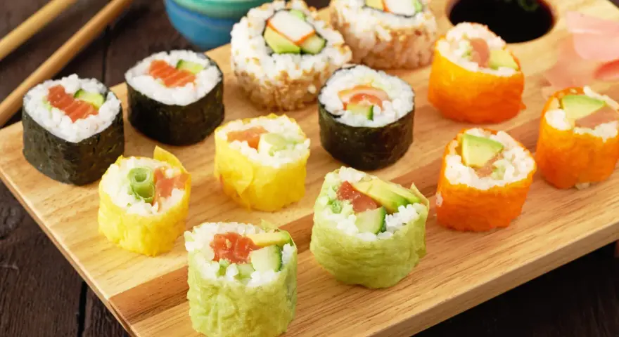 Kolorowe rolki sushi, w tym te z łososiem i awokado, gotowe do zawijania sushi, leżą na drewnianej desce.