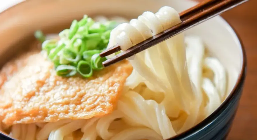 Miseczka z makaronem udon, polana sosem, posypana szczypiorkiem. Pałeczki unoszą porcję grubego makaronu. Pyszne danie, które warto znać pod kątem makaron udon kcal.