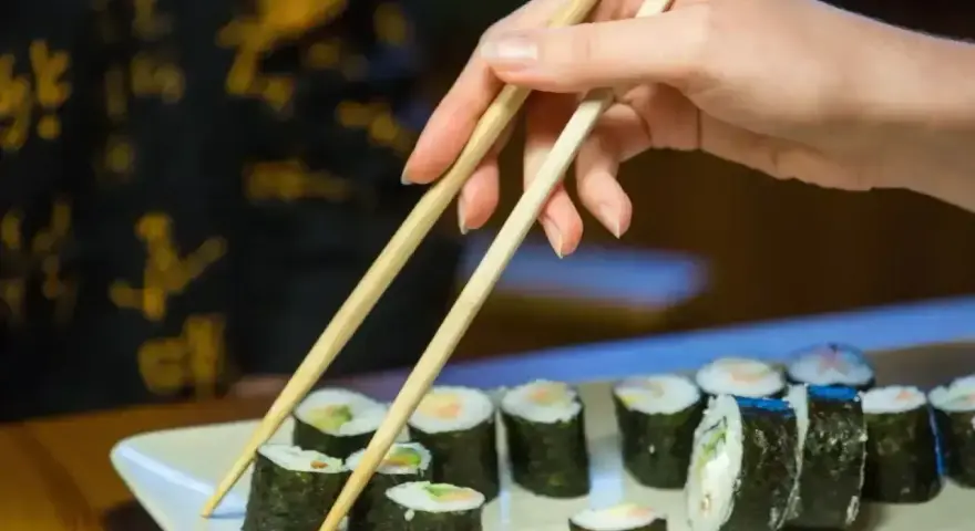 Dłoń podnosi pałeczkami rolkę sushi. Na talerzu ułożone są kawałki sushi, każdy z nich ma około 30 kcal.