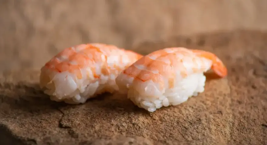 Dwa kawałki sushi z ryżem i różowymi krewetkami do sushi, ułożone na kamiennej powierzchni.