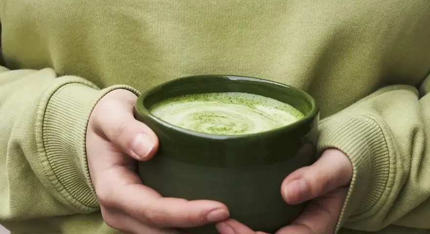 Dłonie trzymające zieloną miseczkę z pianką matcha.