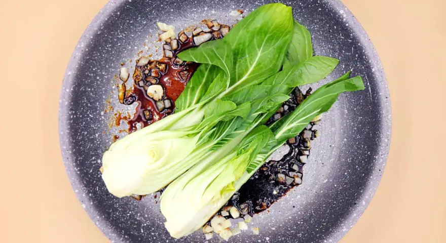 Na patelni smaży się aromatyczny sos z czosnkiem i kawałkami cebuli, a na nim leżą świeże liście sałaty pak choi.