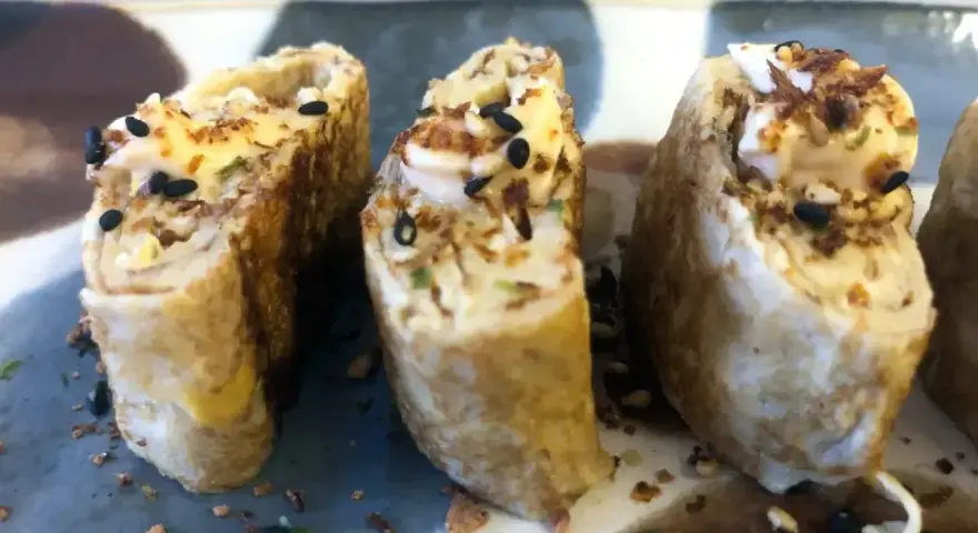 Trzy kawałki sushi z jajkiem, posypane czarnym sezamem i chrupiącymi dodatkami.