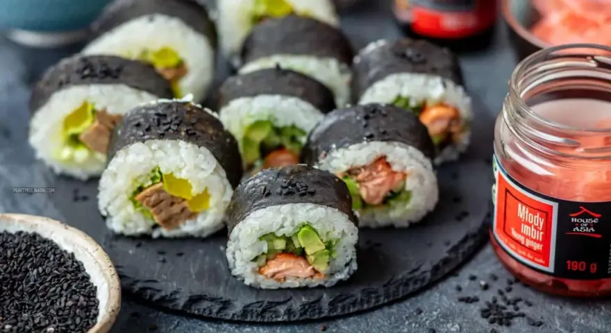 Pyszne sushi z łososiem, awokado i ogórkiem, posypane czarnym sezamem. Obok słoiczek marynowanego imbiru.