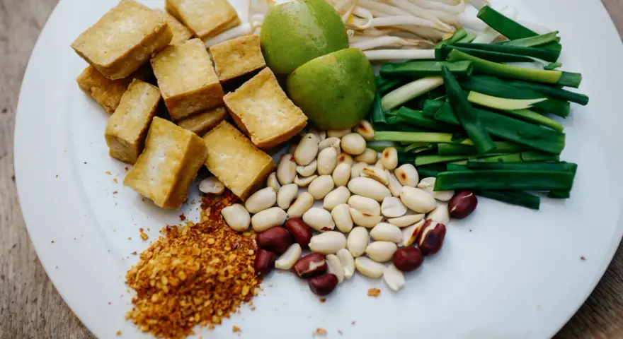 Na talerzu składniki do pad thai: smażone tofu, orzeszki ziemne, szczypiorek, kiełki fasoli mung, limonki i płatki chili. Gotowy sos pad thai czeka!