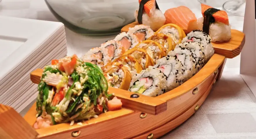 Drewniana łódka pełna sushi, przypominająca o tym, skąd pochodzi sushi. Obok stos białych talerzyków.
