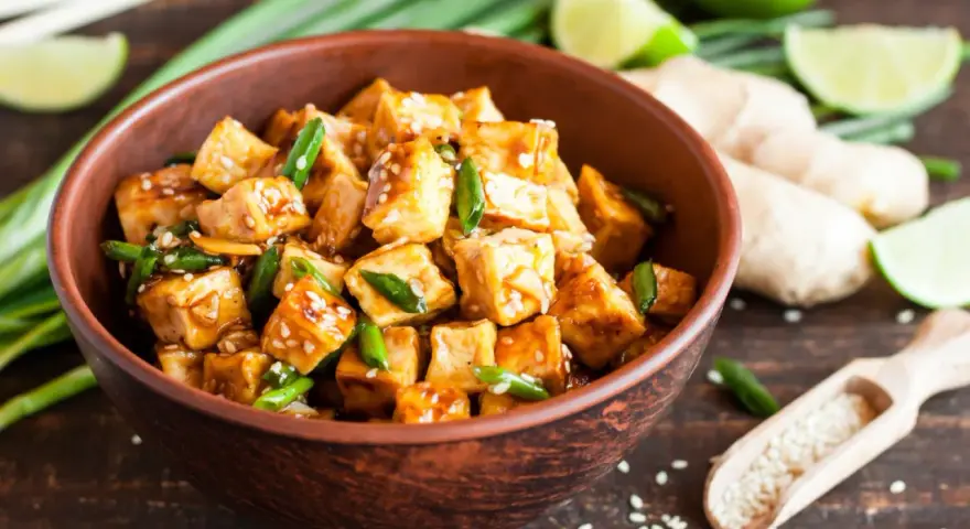 Pokrojone w kostkę, glazurowane tofu z zieloną cebulką i sezamem w misce. Obok limonka, imbir i drewniana łyżka z sezamem.