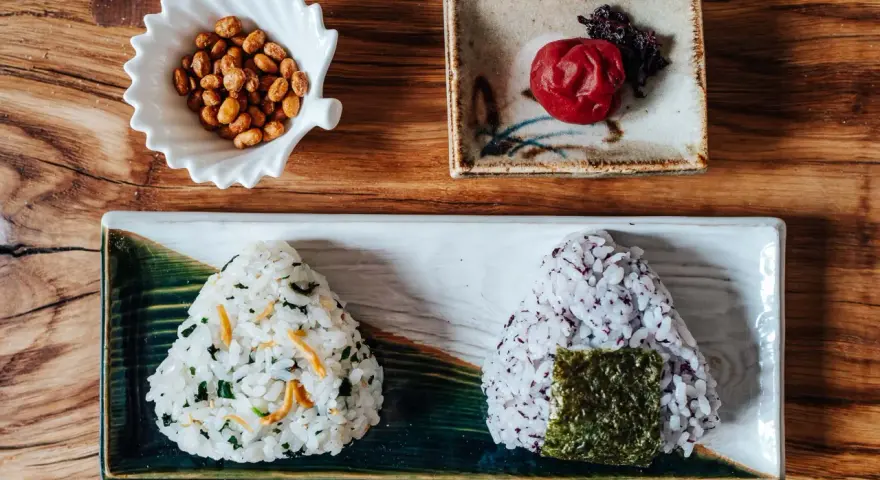 Dwa onigiri z różnym nadzieniem do onigiri: jedno z zielonymi dodatkami i rybkami, drugie z fioletowym ryżem i nori. Obok miseczka orzeszków i marynowana śliwka.