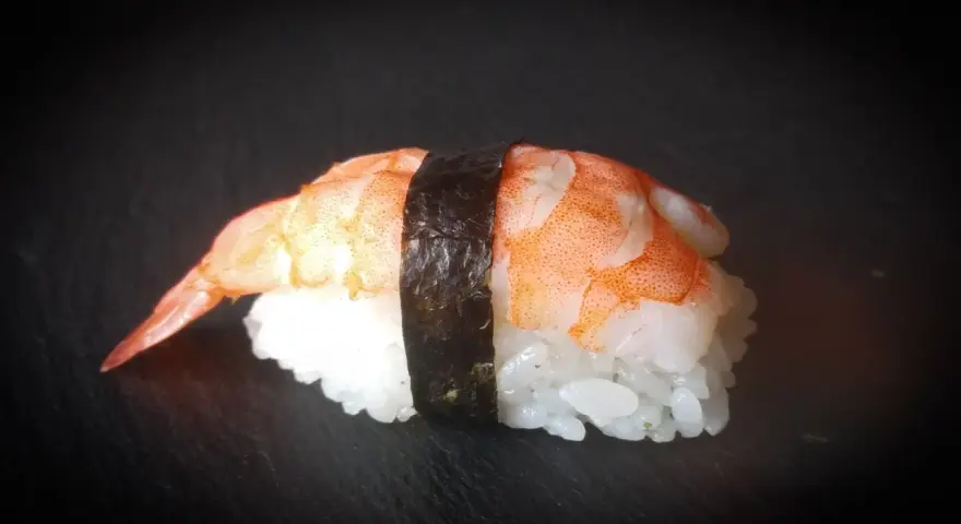 Nigiri ebi, czyli sushi z krewetką, na czarnym tle. Krewetka jest ułożona na ryżu, a całość spięta paskiem nori.