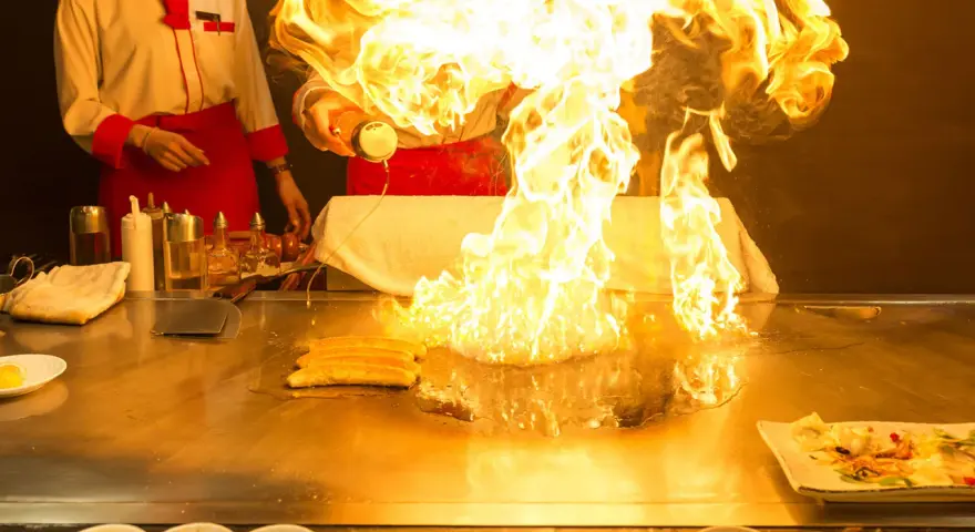 Pokaz teppanyaki z płomieniami buchającymi z płyty grillowej. Kucharz w czerwonym fartuchu dodaje składniki, tworząc widowiskowy spektakl.