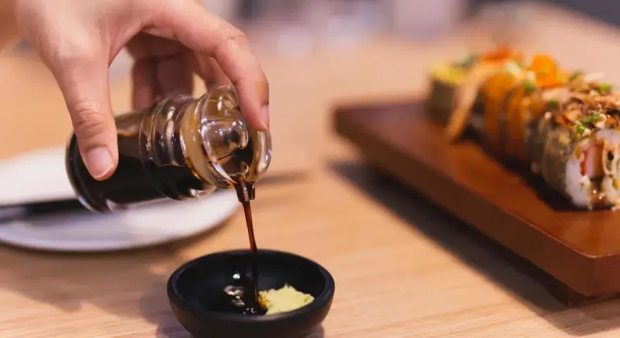 Dłoń nalewa sos sojowy bezglutenowy do miseczki z wasabi. Obok leży sushi.