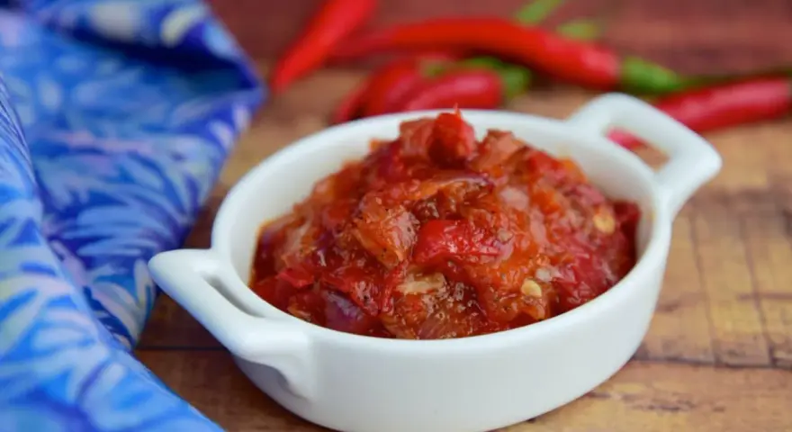 Pikantny sos sambal oelek w białej miseczce, obok czerwonych papryczek chili i niebieskiej tkaniny.