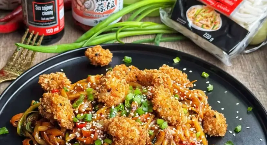 Pyszny **udon z kurczakiem i warzywami**, posypany sezamem i szczypiorkiem. Obok sosy i opakowanie makaronu.