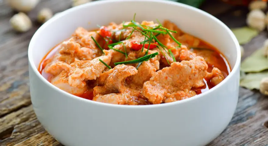Aromaticzne curry z wieprzowiną w białej misce, ozdobione zieloną szczypiorkiem i czerwonymi papryczkami chili.