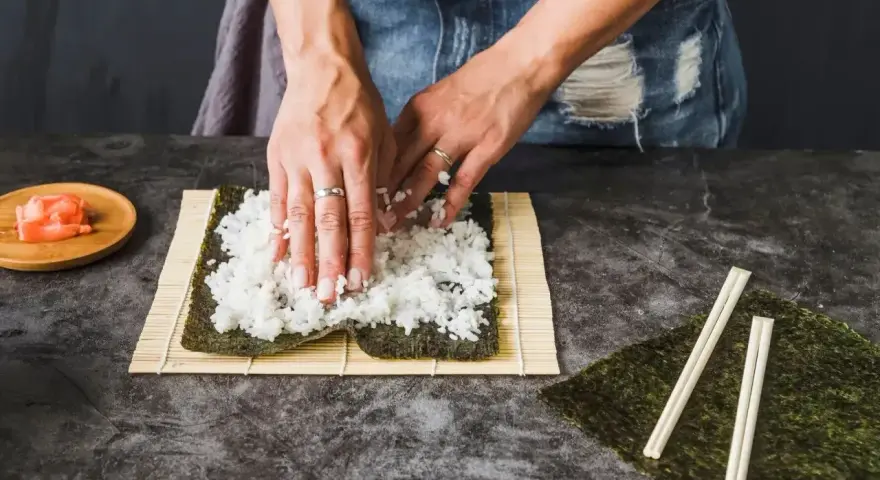 Dłonie rozprowadzają ryż na nori. Obok leżą pałeczki i marynowany imbir. To klucz do idealnego ryżu do sushi, który potrzebuje odrobiny octu.