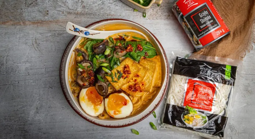 Szybki ramen przepis: miska pełna aromatycznego bulionu z grzybami shiitake, tofu, jajkiem i szczypiorkiem. Obok opakowanie makaronu i grzybów.