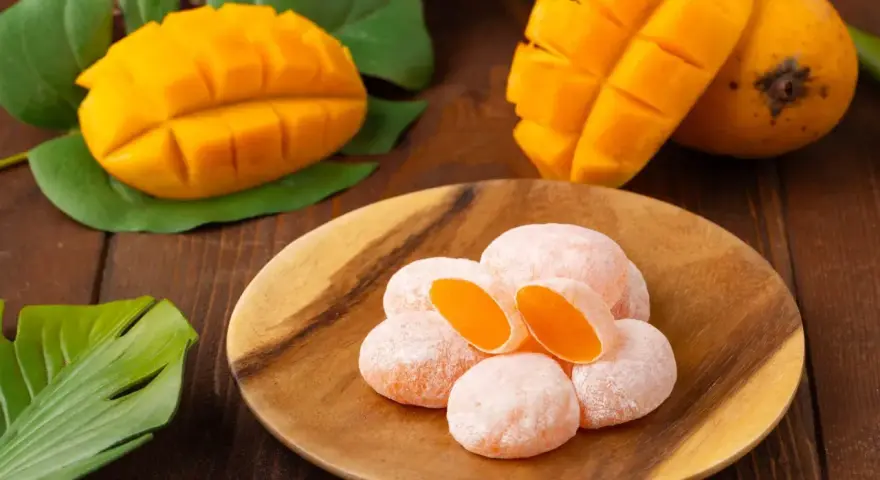 Słodkie mochi o smaku mango, ułożone na drewnianym talerzu, obok świeżych owoców i liści.