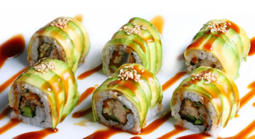 Sześć kawałków sushi dragon roll, udekorowanych plastrami awokado, sezamem i sosem.