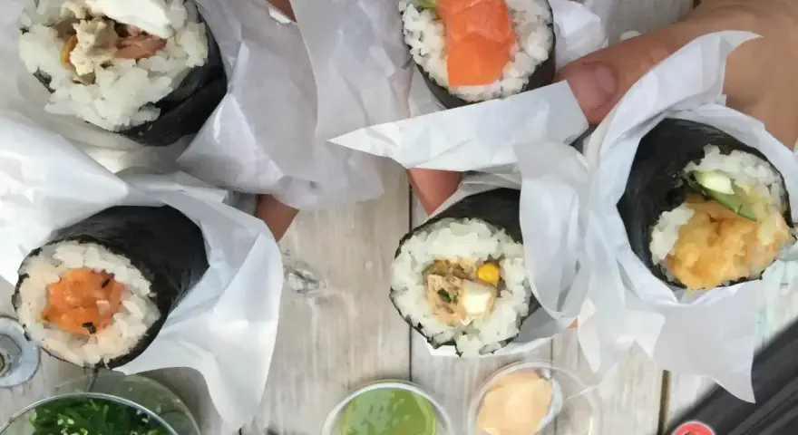 Ręce trzymają cztery pyszne temaki zawinięte w papier. Widać różne nadzienia: łososia, awokado, warzywa i rybę.